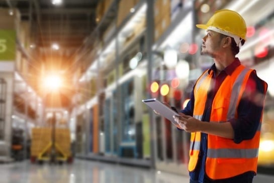 A man standing in the warehouse holding a pad, wearing a safety helmet and a safety vest. Shelves and a moving forklift in the background.